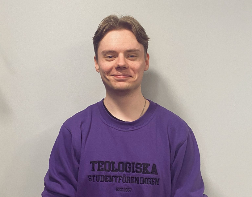 Jonas Väänänen tittar rakt in i kameran. Han har på sig en lilafärgad tröja med texten Teologiska studentföreningen.